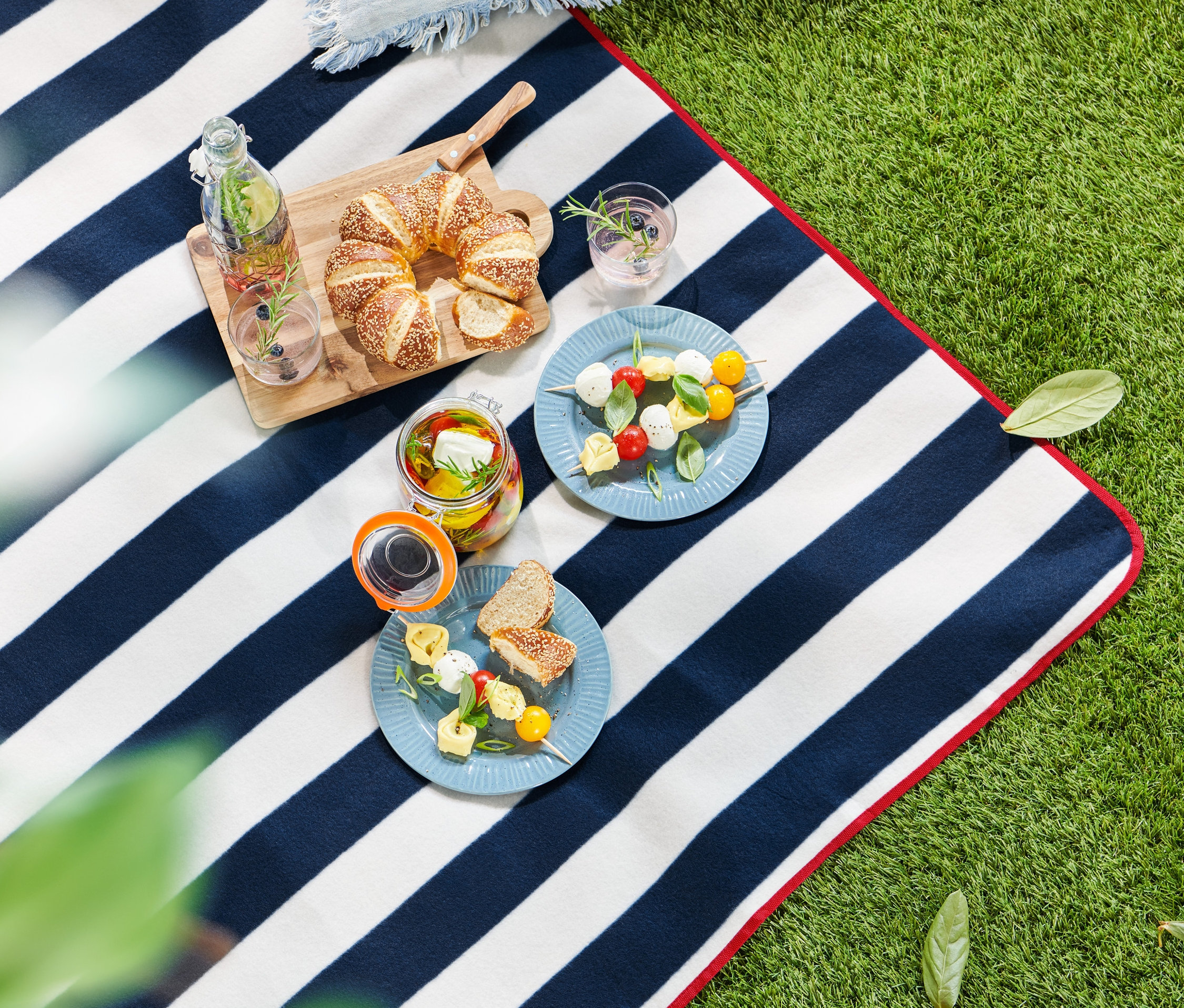 Çim üzerinde mavi ve beyaz şeritli küçük bir piknik örtüsünde yiyecek ve içecek.