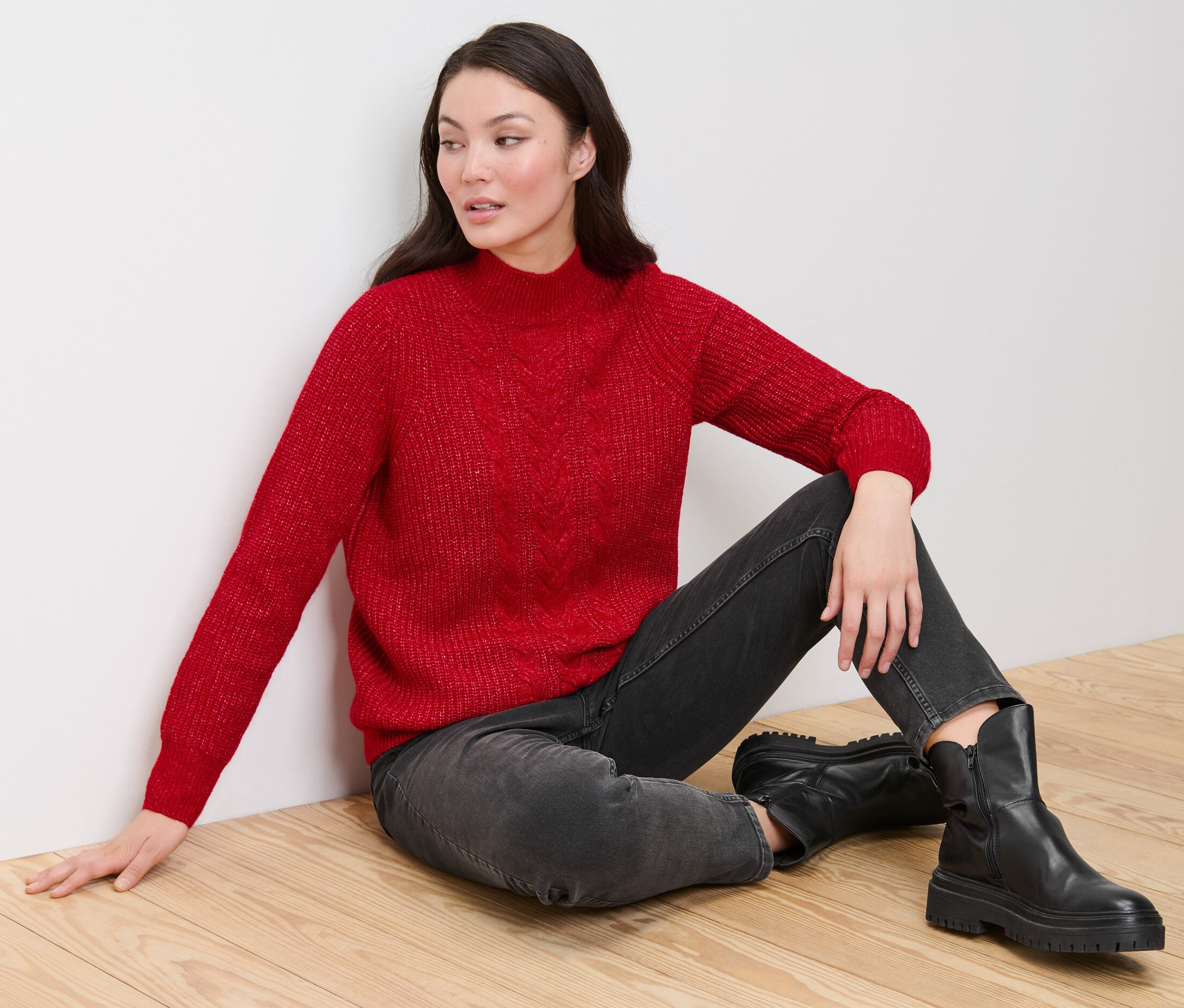 Ahşap zeminde oturan bir kadın, kırmızı, saç örgüsü motifli kalın örgü kazak, siyah kot pantolon ve siyah botlar giyiyor.