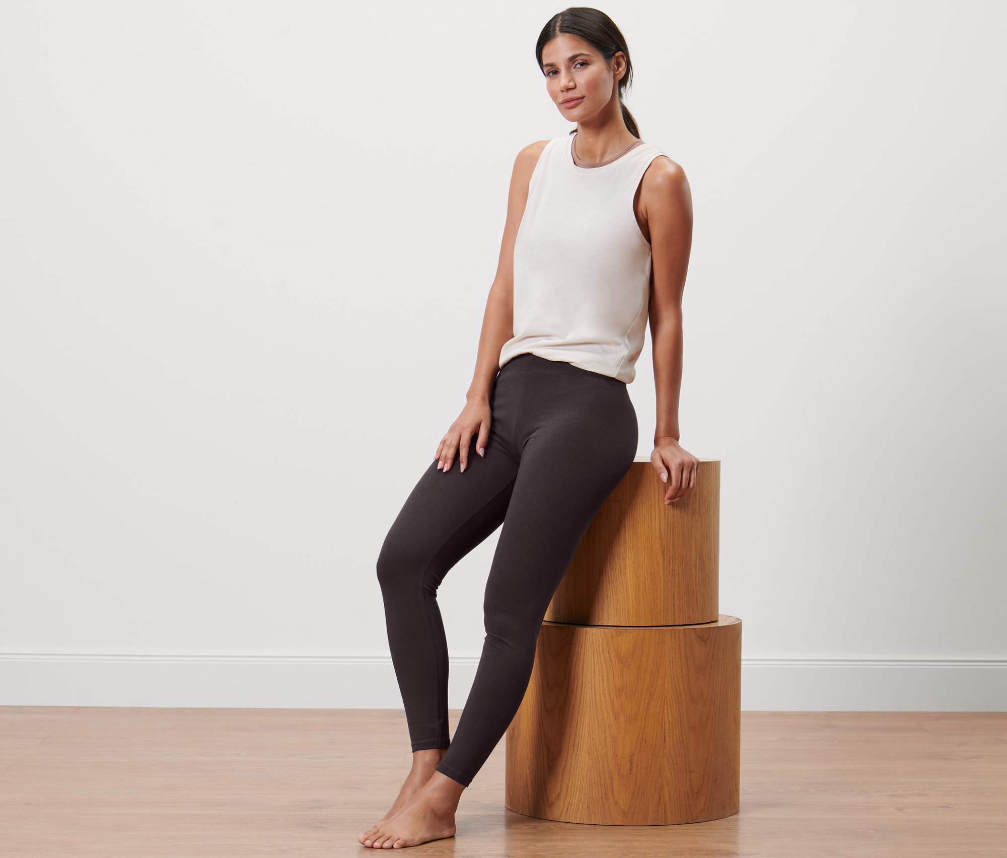 Kadın ahşap silindirlerin üzerinde oturuyor, spor taytı ve atlet giyiyor.