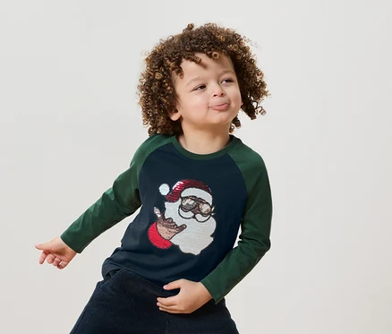 Kıvırcık saçlı bir çocuk, üzerinde Noel Baba çizimi olan mavi-yeşil bir tişört giyiyor.