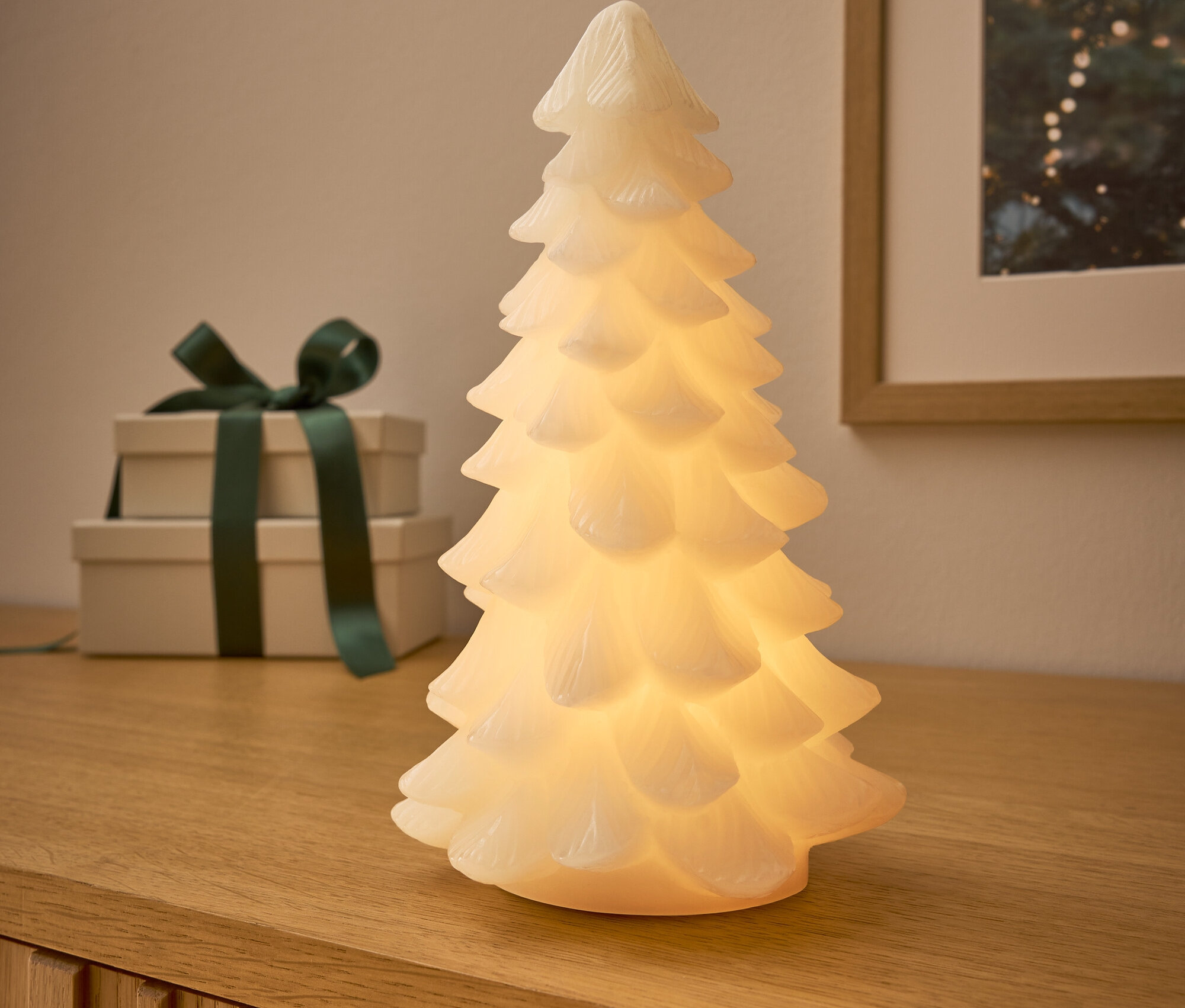 Ahşap bir komodinin üzerinde parlayan bir ağaç duruyor, arkasında koyu yeşil kurdeleli iki hediye ve Noel temalı bir resim var.