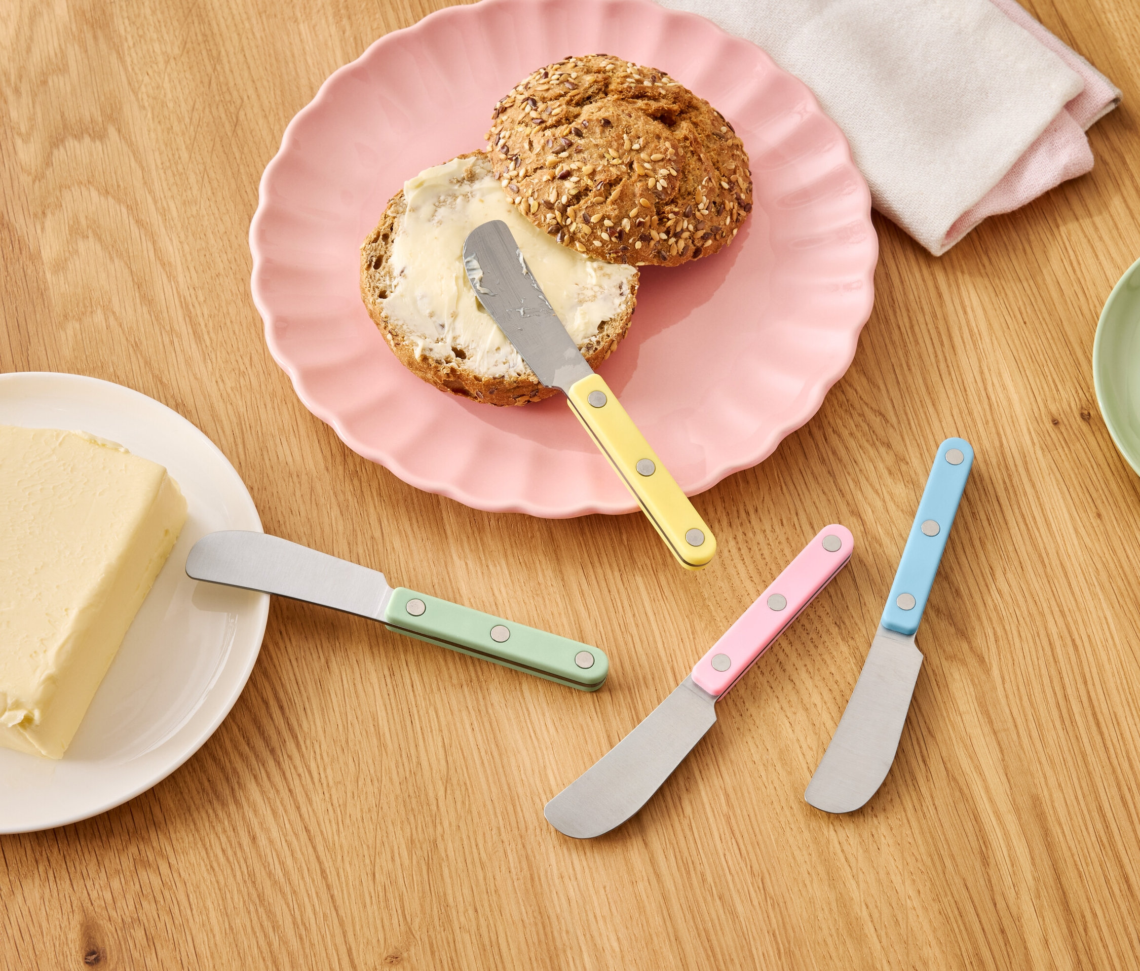 Tahta bir masa üzerinde pembe bir tabakta tereyağlı bir rulo, bir parça tereyağı ve renkli bıçaklar.