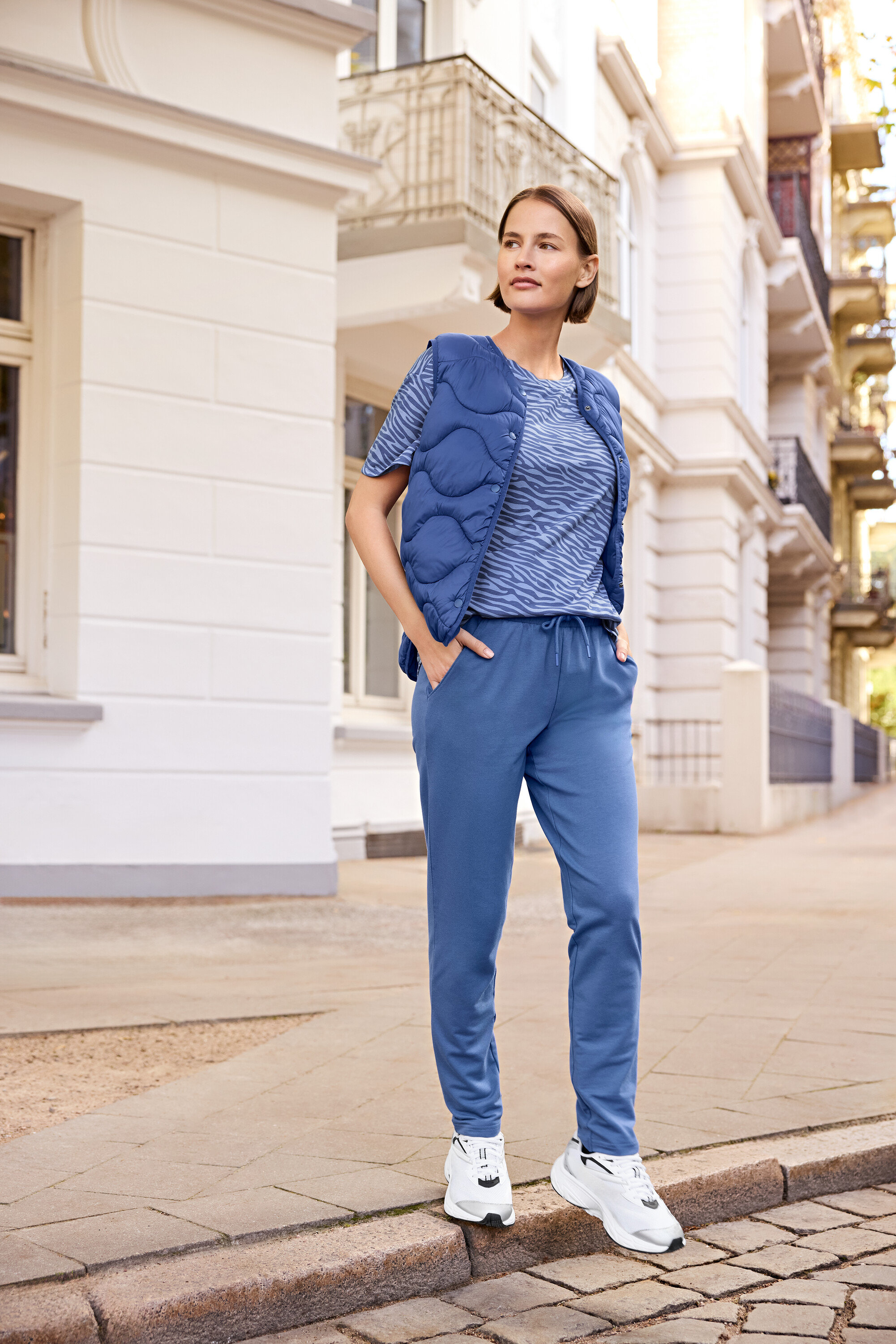 Kadın kaldırımda duruyor, üzerinde mavi yelek, zebra desenli mavi tişört ve mavi eşofman altı var.