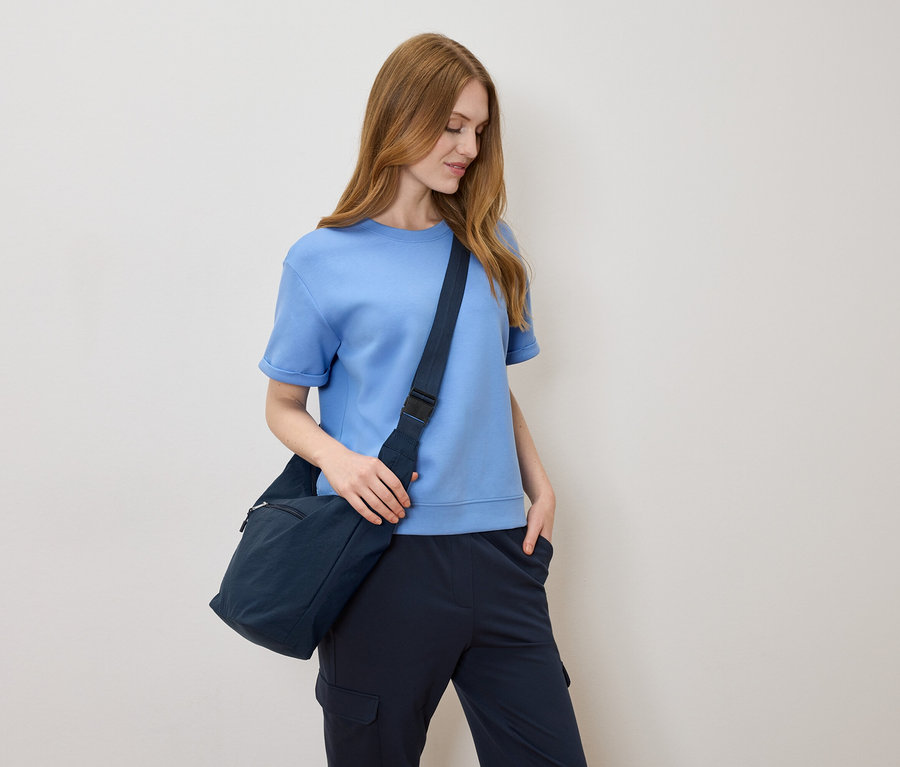 Açık kahverengi saçlı, açık mavi tişörtlü ve krem rengi bir arka planda lacivert omuz çantalı kadın.