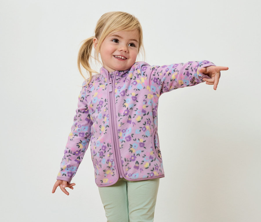 Sarışın örgülü, çiçek desenli mor polar ceket ve açık yeşil pantolon giyen gülümseyen bir kız çocuğu parmağıyla işaret ediyor.