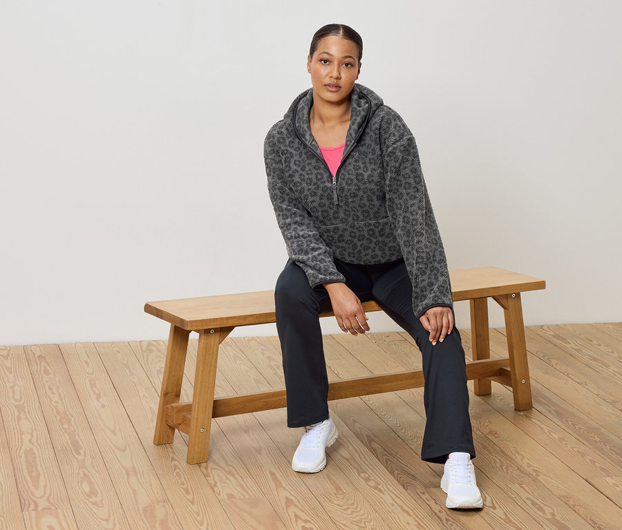 Kadın ahşap bir bankta oturuyor, leopar desenli gri bir polar kazak, pembe bir üst, koyu renk pantolon ve beyaz spor ayakkabılar giyiyor.