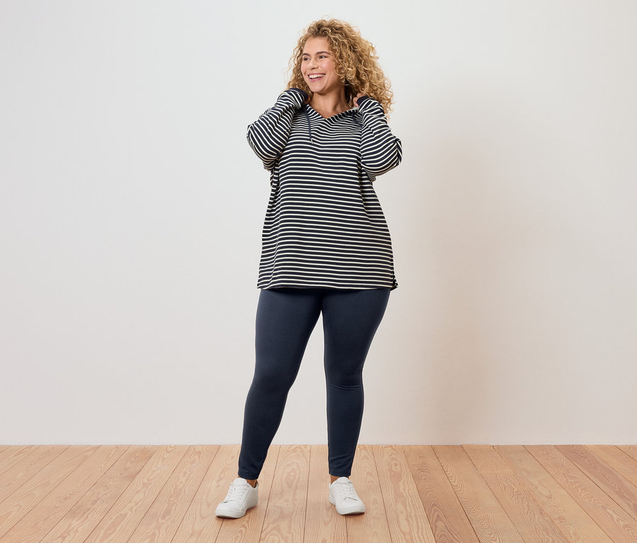 Kıvırcık saçlı bir kadın, çizgili bir üst, tayt ve beyaz spor ayakkabı giyiyor.