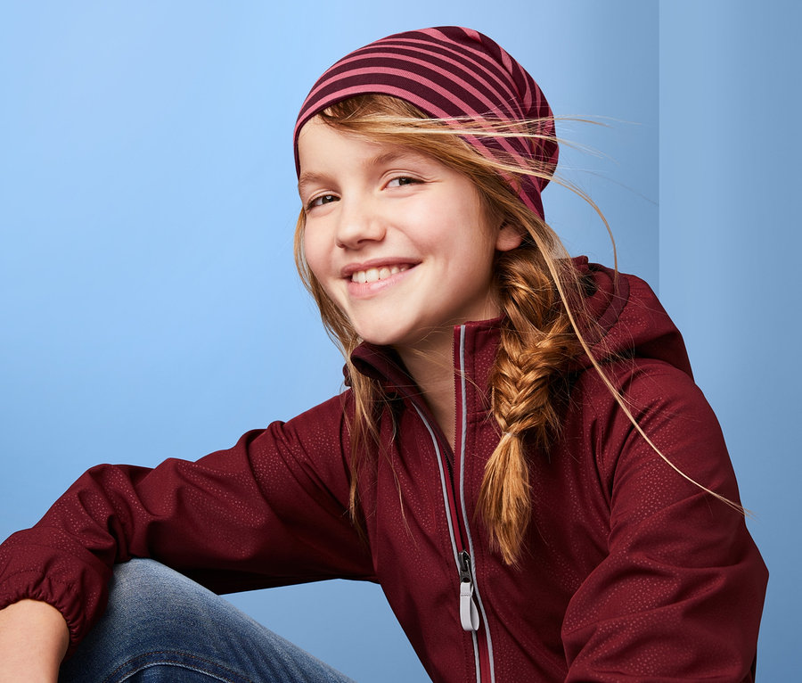 Gülümseyen kız bordo çizgili Organik Pamuklu Çift Taraflı Bere ve bordo bir ceket giyiyor.