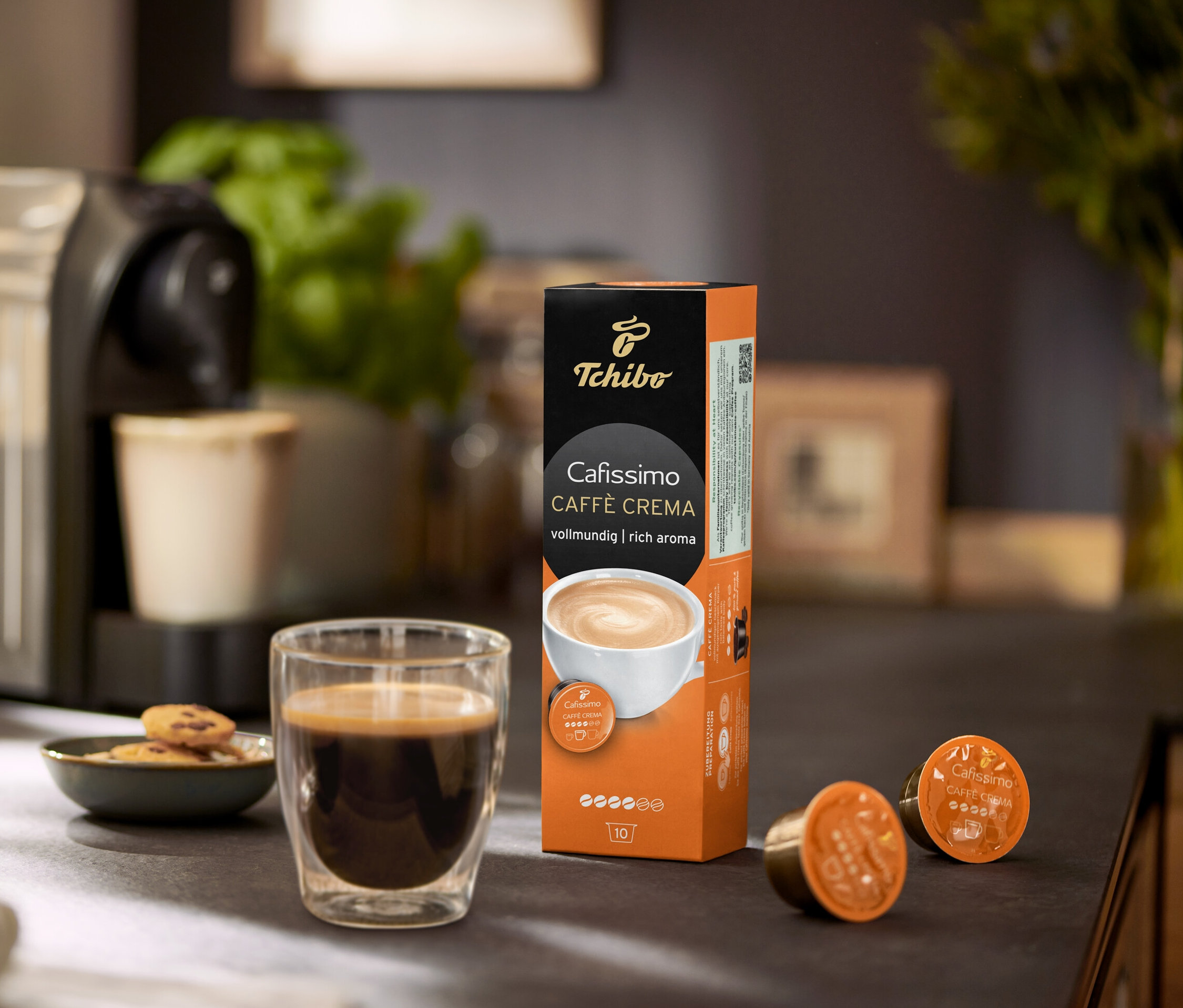 Kahve makinesi, bir fincan kahve, kurabiyeler ve bir paket Cafissimo Caffe Crema kapsülü ile birlikte.