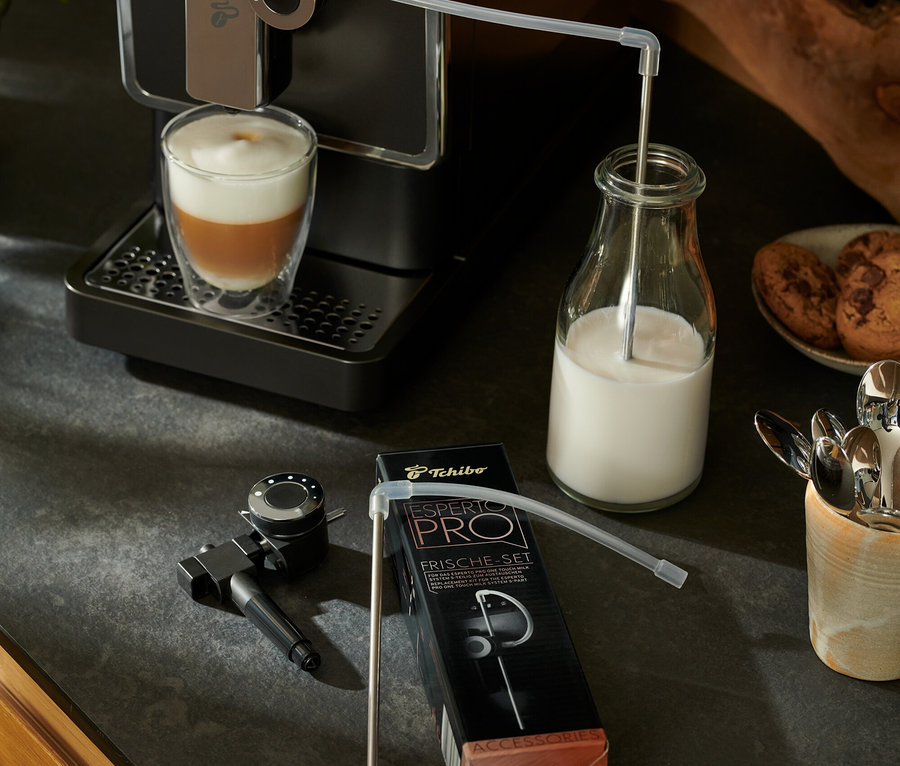 Antrasit renkli Tchibo »Esperto Pro« tam otomatik kahve makinesi bir içecek hazırlıyor. Süt bir hortum aracılığıyla cam bir şişeye akıyor. Yanında bir Esperto Pro &amp; Esperto2 Milk yedek seti duruyor.
