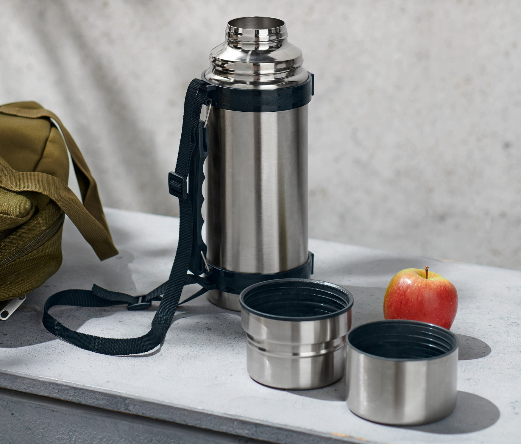 Gümüş renkli, iki bardaklı termos, zeytin yeşili bir çanta ve bir elma gri bir yüzey üzerinde duruyor.