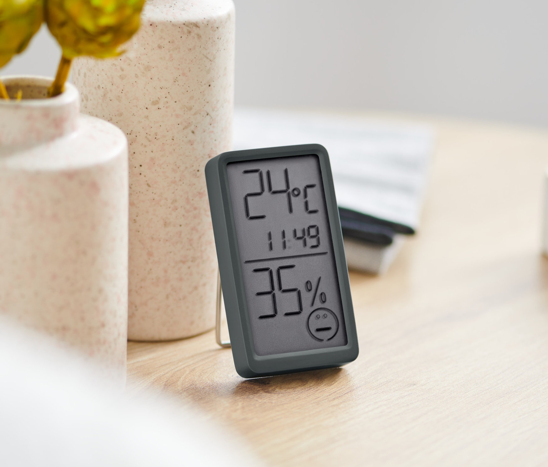 Masanın üzerinde 2 Adet Oda Termometresi, çiçek vazoları ve bir kitap var.