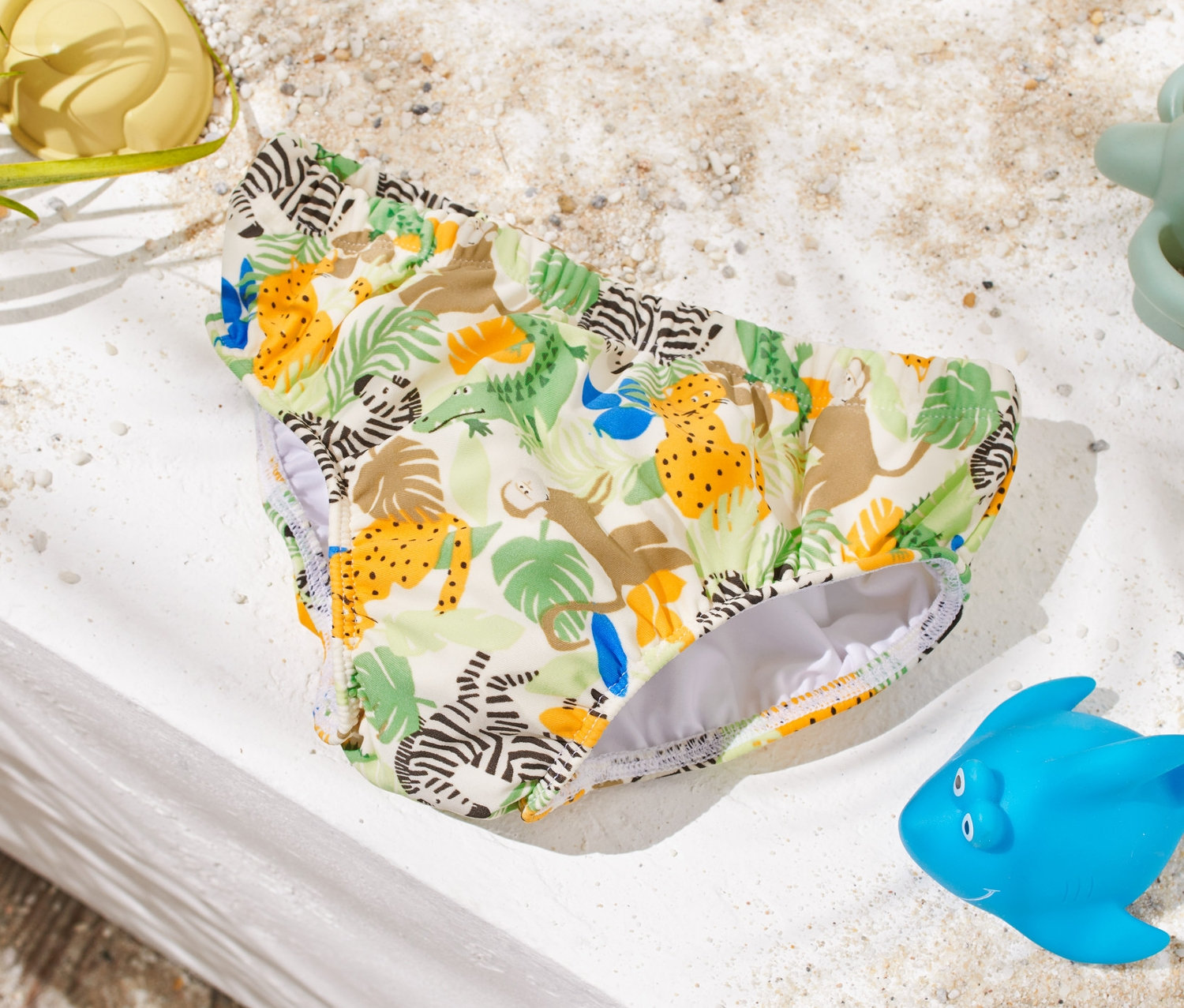 Hayvan desenli bebek külot mayo, kum oyuncaklarının yanında beyaz bir yüzeyde duruyor.