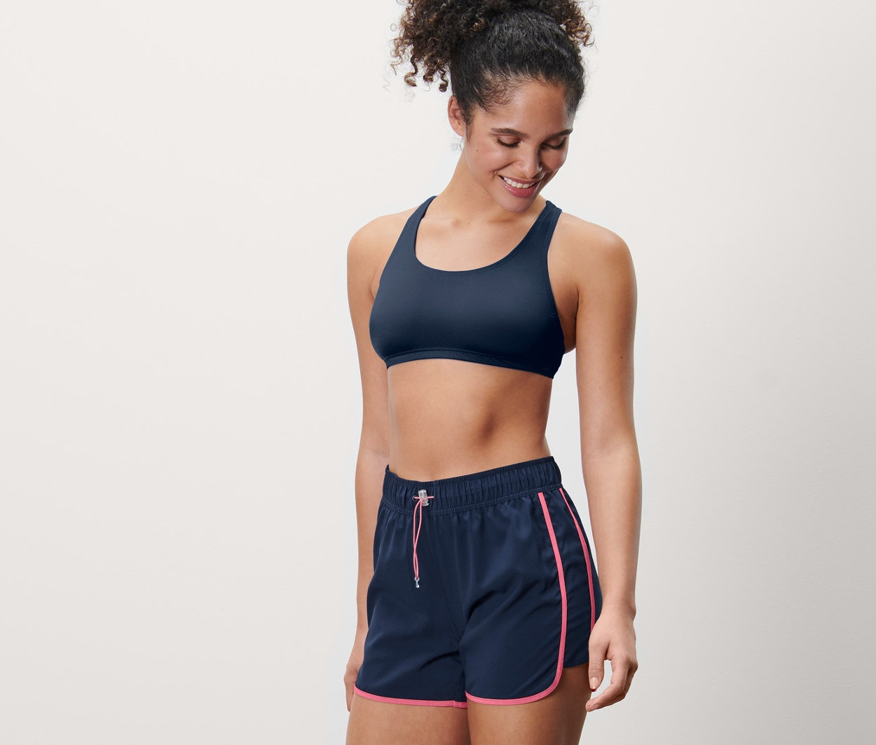 Gülümseyen bir kadın şort mayo ve sporcu sütyeni giyiyor.