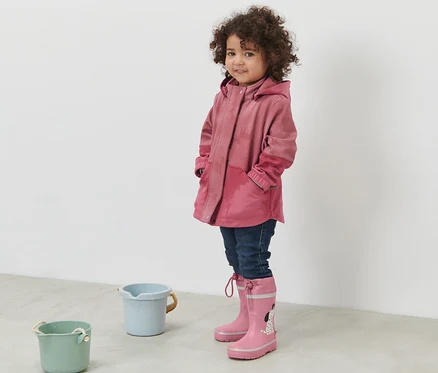 Ayakta duran bir kız, yağmurda renk değiştiren pembe çocuk softshell montu ve pembe lastik çizme giyiyor.