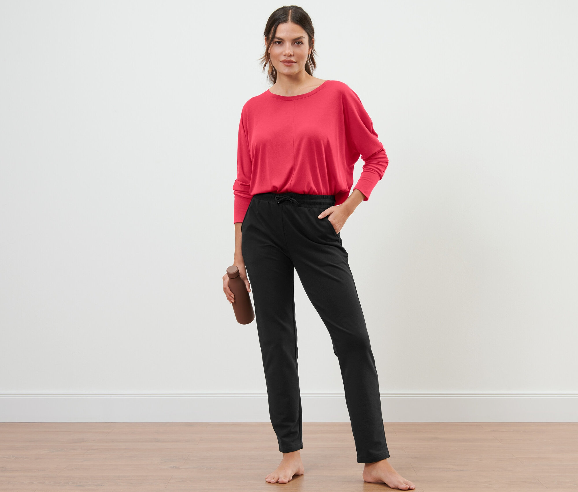 Kadın ayakta duruyor ve bir su şişesi tutuyor, üzerinde siyah bir eşofman altı var.