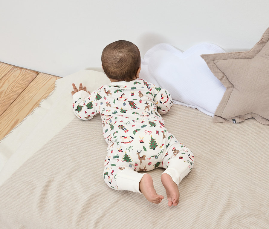 Bebek, Noel desenli bir battaniyenin üzerinde, beyaz ve bej yastıkların yanında yüzüstü yatıyor.