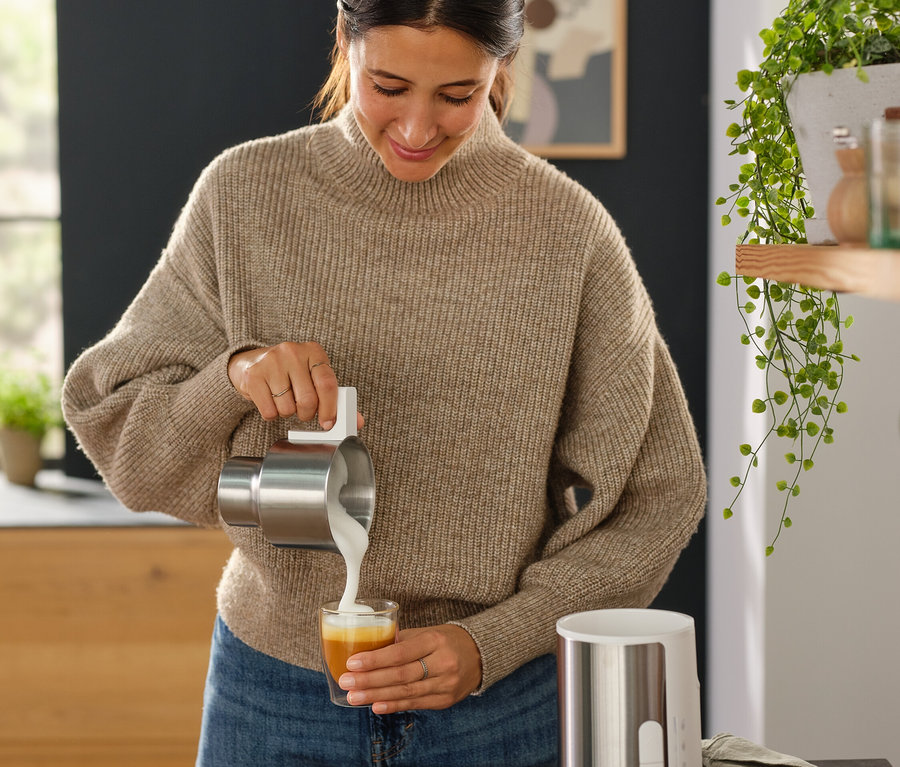 Kadın, paslanmaz çelik bir kaptan siyah bir indüksiyonlu süt köpürtücünün yanındaki bir bardağa süt döküyor.