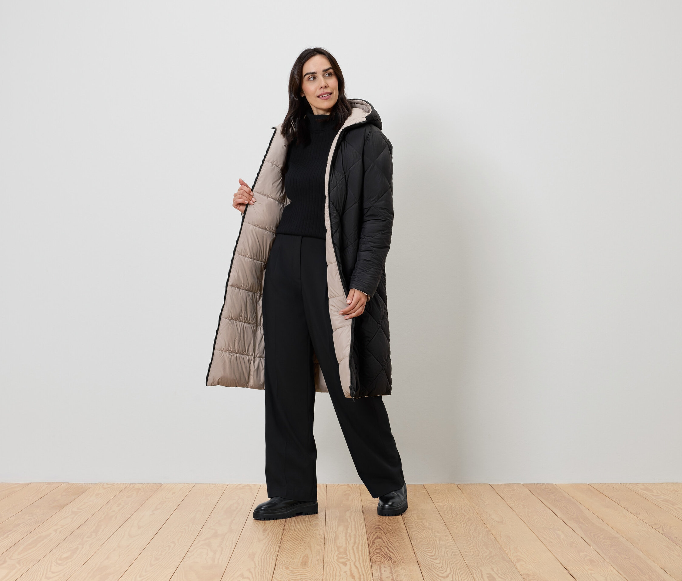 Kadın bej astarlı siyah bir palto, siyah balıkçı yaka ve siyah pantolonla poz veriyor.