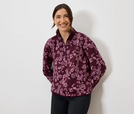 Gülümseyen bir kadın, bordo çiçekli polar bir sweatshirt ve koyu renk pantolon giyiyor.
