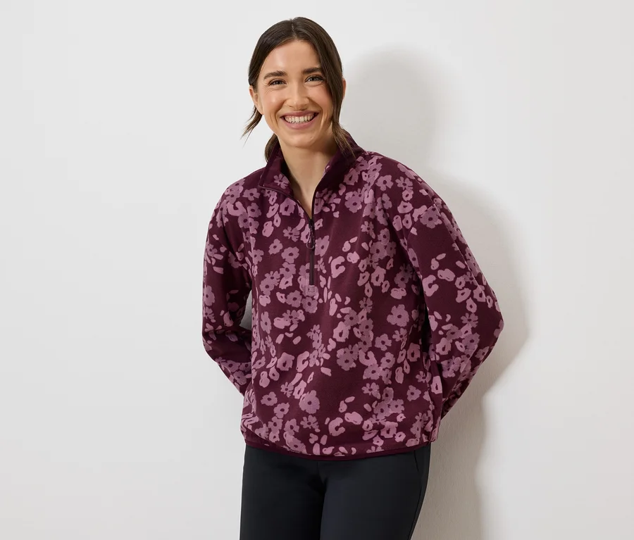 Gülümseyen bir kadın, bordo çiçekli polar bir sweatshirt ve koyu renk pantolon giyiyor.