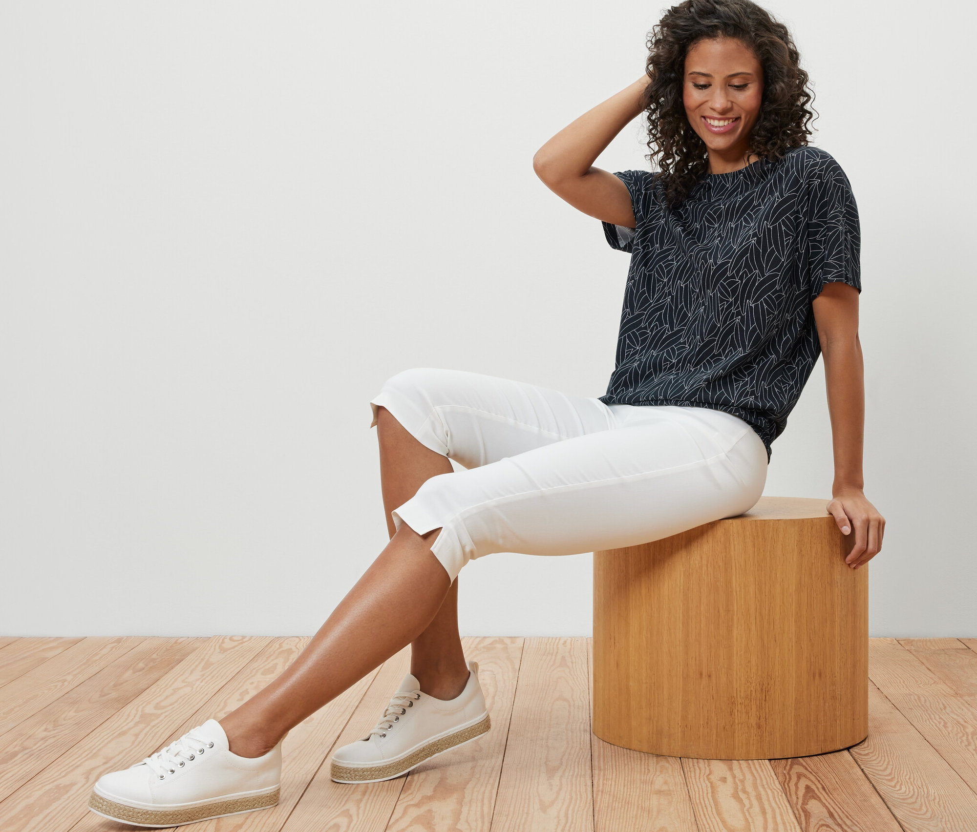 Bir kadın taburede oturuyor ve lacivert, düşük omuzlu bir tişört, beyaz kapri pantolon ve spor ayakkabı giyiyor.