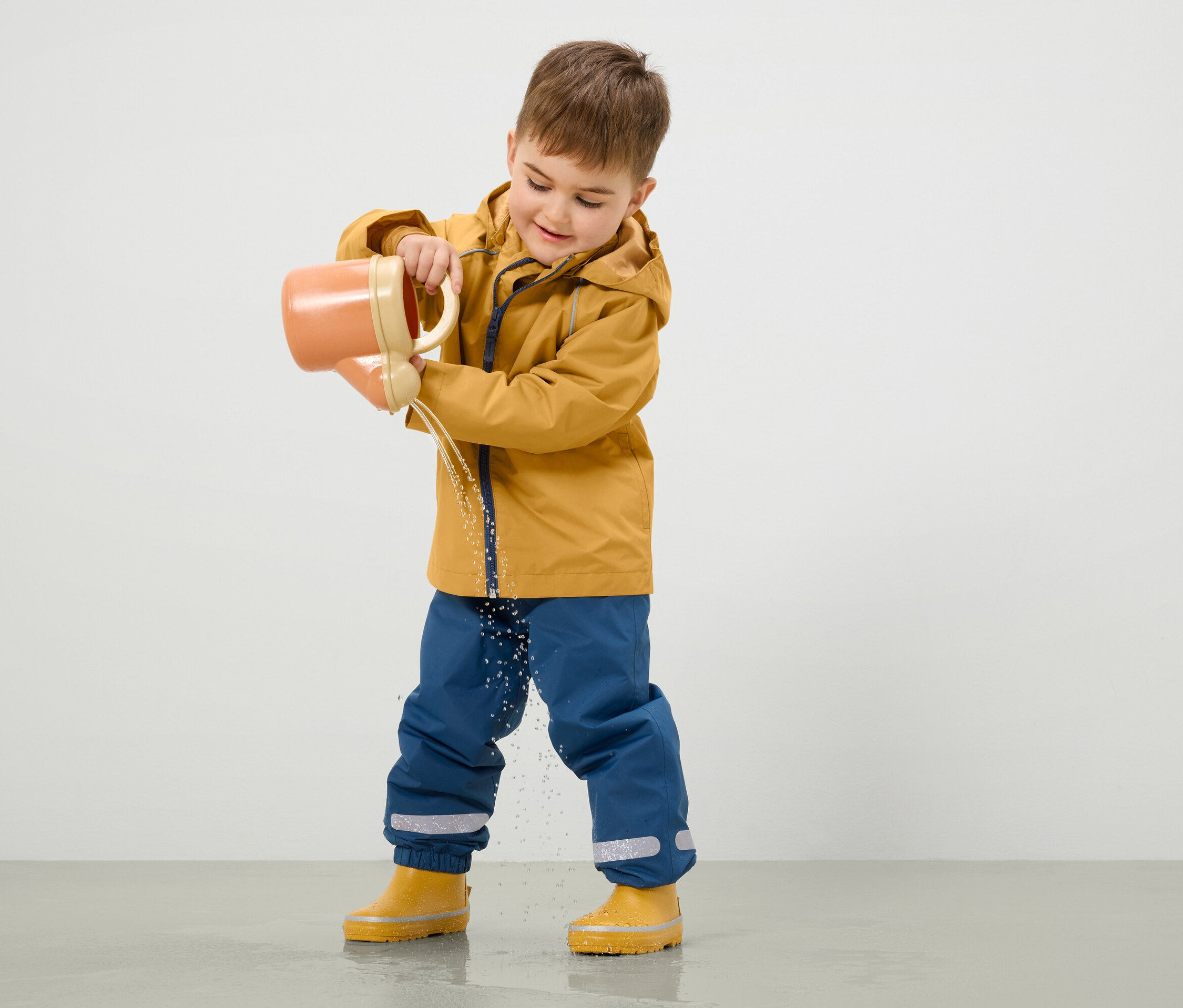 Sarı montlu ve mavi pantolonlu bir çocuk, turuncu bir sulama kabından yere su döküyor.