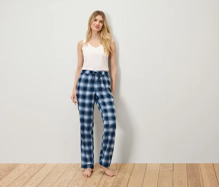 Bir kadın ahşap bir zeminde duruyor, beyaz bir atlet ve mavi kareli bir pijama pantolonu giyiyor.