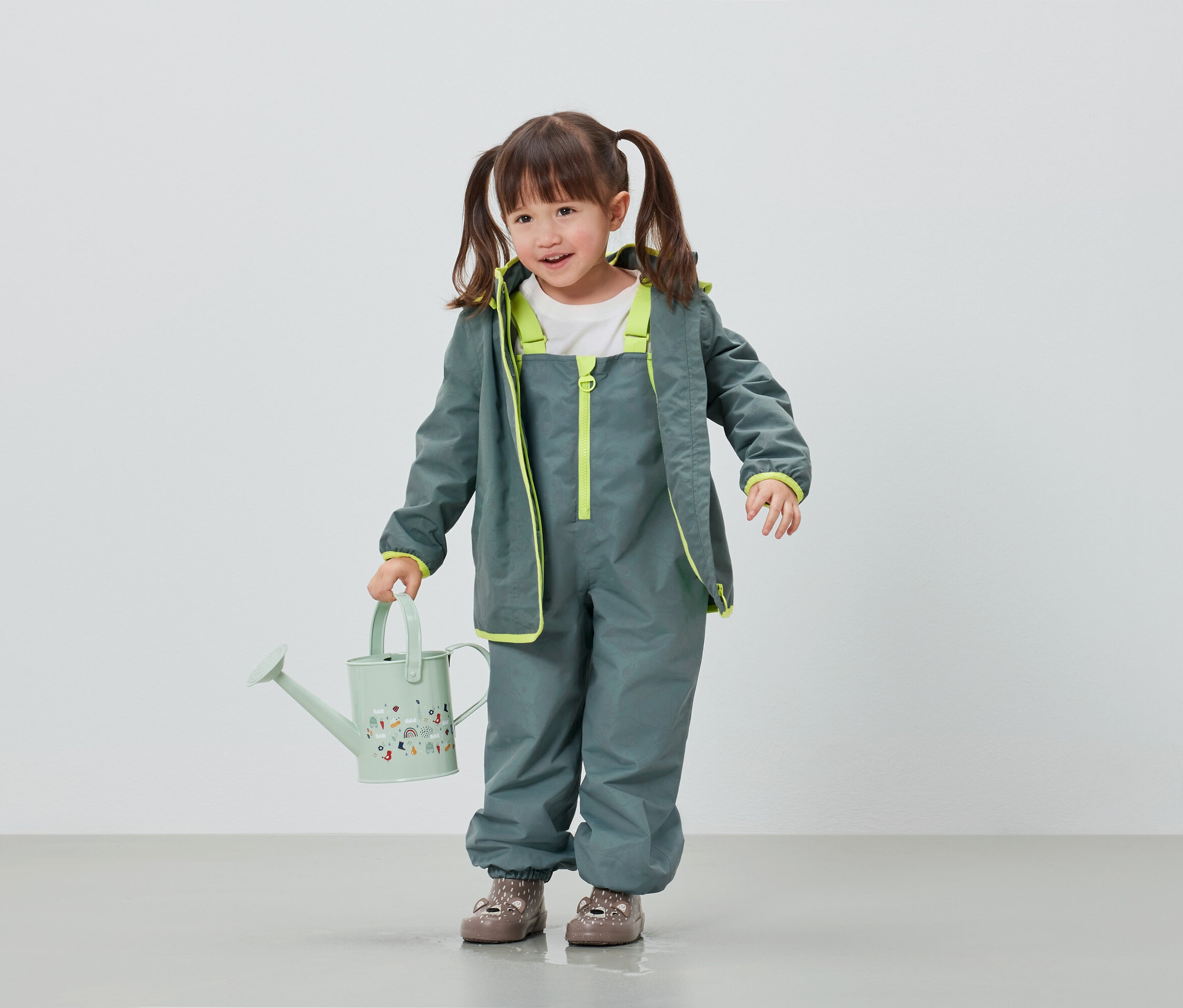 Gri yağmurluk ve pantolon giyen kız çocuğu açık yeşil sulama kabı tutuyor.
