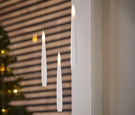 İplerde asılı duran üç adet beyaz, elektronik mum. Arka planda bir Noel ağacı ve ahşap bir duvar var.