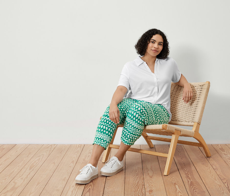 Bir kadın sandalyede oturuyor, üzerinde baskılı 3/4 jersey pantolon ve beyaz düğmeli bluz var.