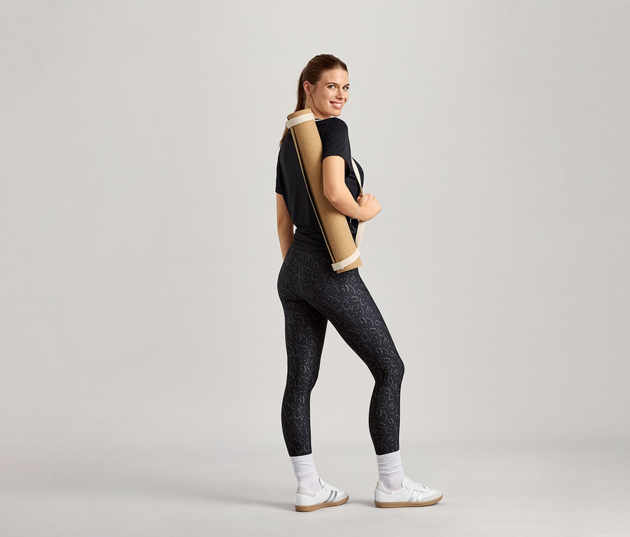 Gülümseyen kadın omzunda yoga matı, üzerinde siyah spor tişört var.