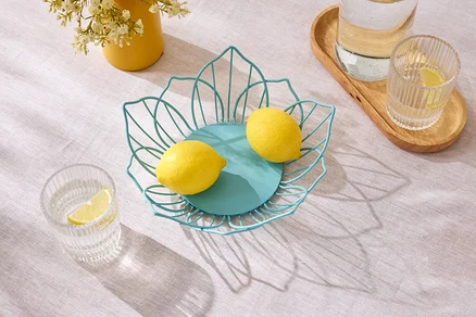 Sarı çiçekli sarı bir vazo ve su bardaklarının yanında bir masanın üzerinde mavi bir tel kabın içinde iki limon duruyor.