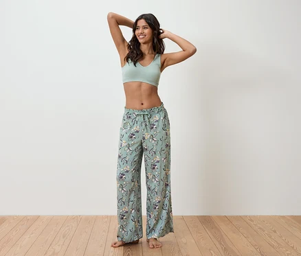 Açık yeşil spor sütyeni ve çiçekli pijama altı giyen bir kadın duvarın önünde duruyor.