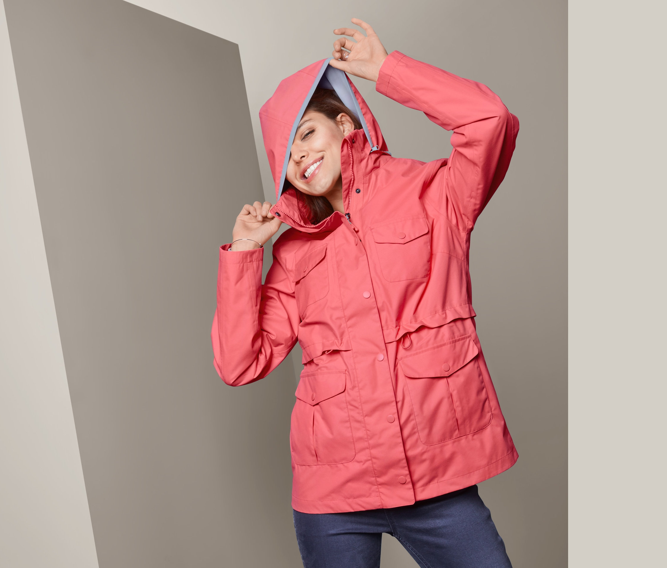 Gülümseyen bir kadın, Mercan Rengi Fonksiyonel Yağmur Montu'nun kapüşonunu başına geçiriyor.