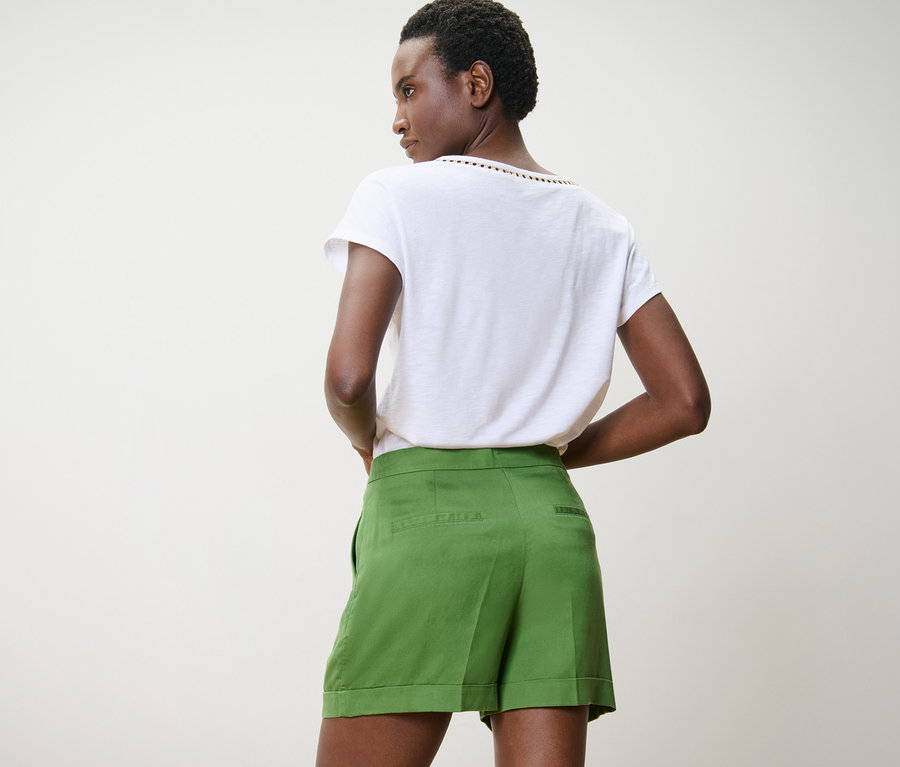 Esmer tenli bir kadın yeşil keten şort ve beyaz tişörtle arkadan poz veriyor.