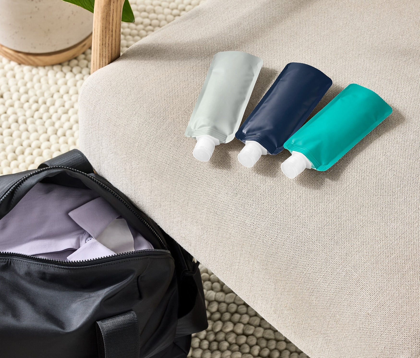 Bej rengi bir koltukta, siyah bir spor çantasının ve saksı bitkisinin yanında üç adet tekrar doldurulabilir yolculuk şişesi duruyor.