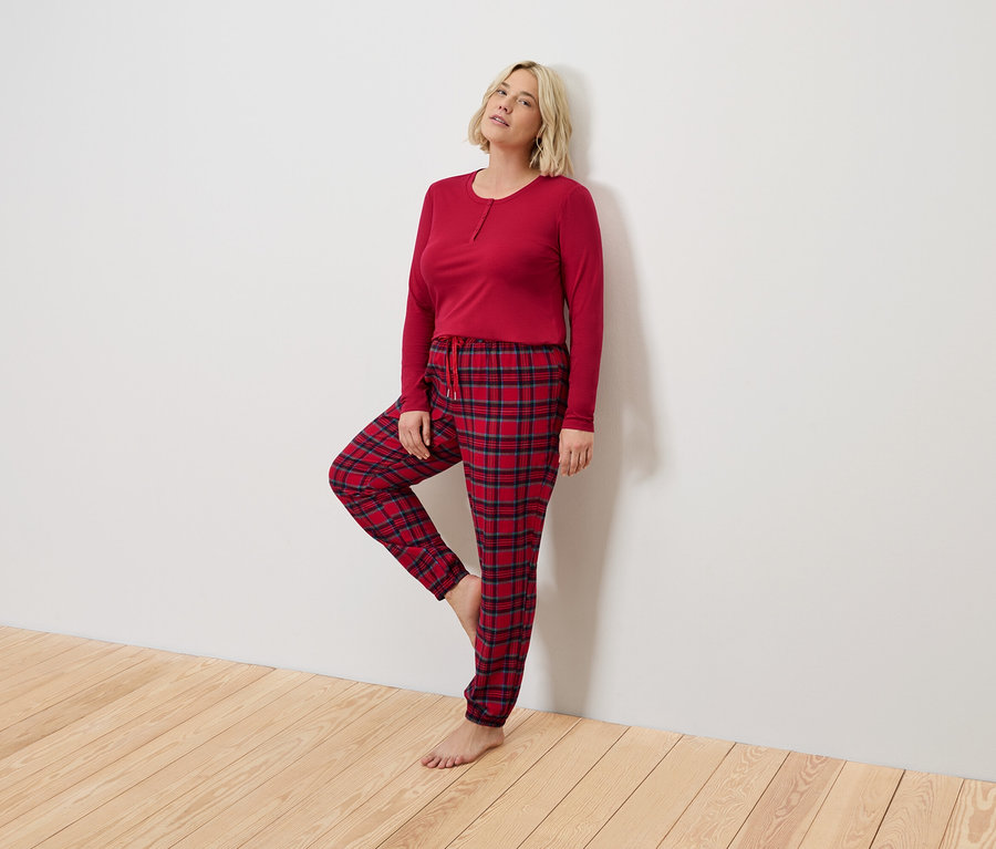 Sarışın bir kadın kırmızı pijama ve ekose desenli pantolonla beyaz bir duvara yaslanmış.