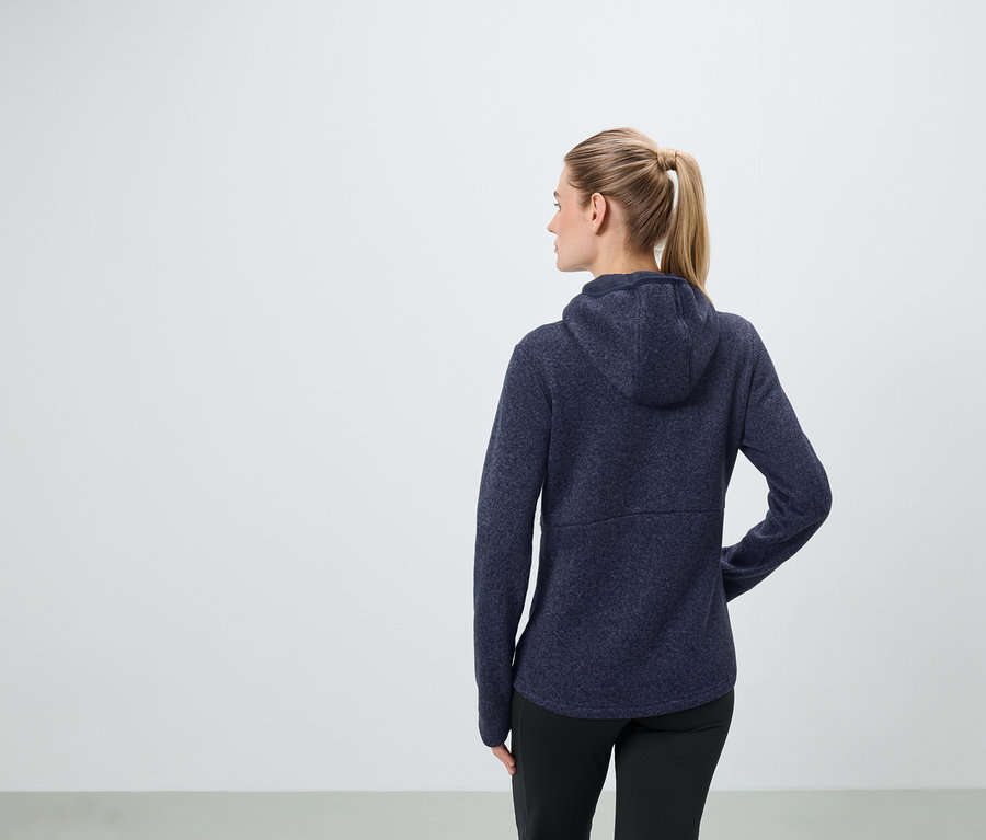Sarışın saçları at kuyruğu şeklinde toplanmış bir kadın, lacivert kapüşonlu bir sweatshirt ve siyah pantolon giyiyor.