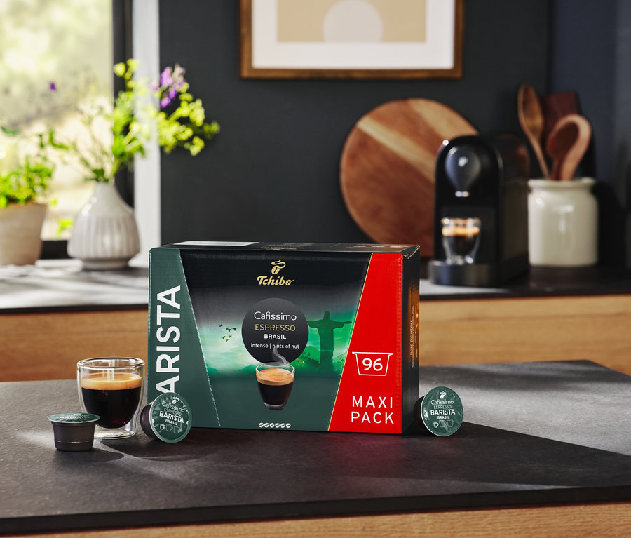 Bir masa üzerinde bir paket Espresso Brasil kapsülü, bir bardak espresso ve bir kahve makinesi bulunmaktadır.