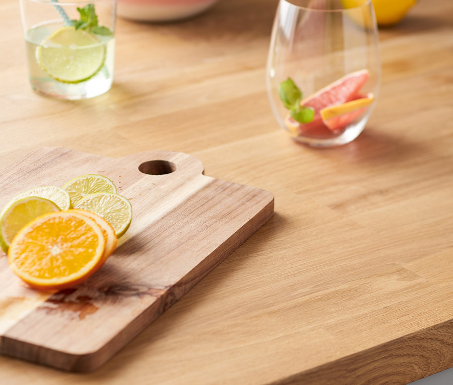 Ahşap kesme tahtası üzerinde portakal ve misket limonu dilimleri, yanında içecek bardağı ve meyve kasesi.