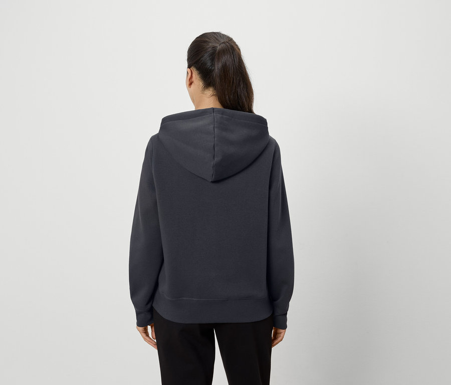 Arkası dönük duran bir kadın, koyu gri kapüşonlu bir sweatshirt ve siyah pantolon giyiyor.