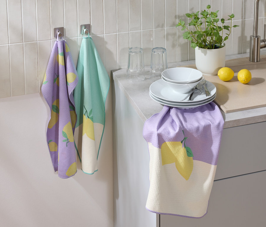 Duvarda limon desenli mor ve yeşil bir mutfak bezi asılı duruyor. Tezgahta tabaklar, bir kase, bardaklar, çatal bıçak takımı, bir limon ve bir çiçek var.