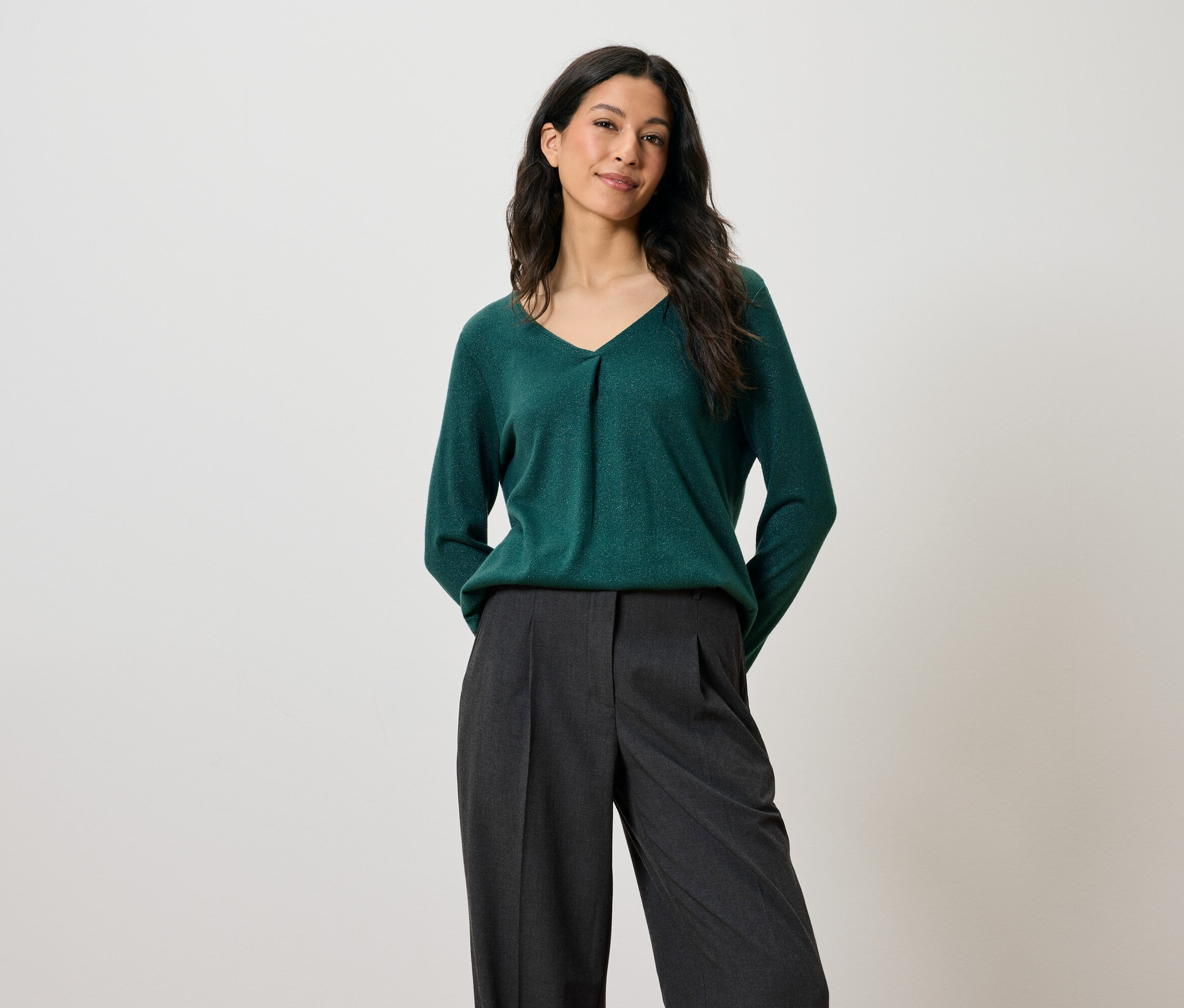 Koyu saçlı bir kadın yeşil bir kazak ve gri pantolonla poz veriyor.