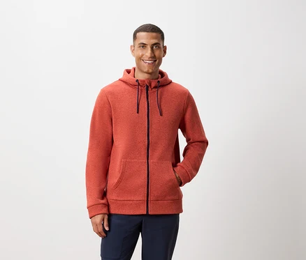 Beyaz bir arka planda poz veren, turuncu fermuarlı kapüşonlu sweatshirt ve lacivert pantolon giyen bir adam.