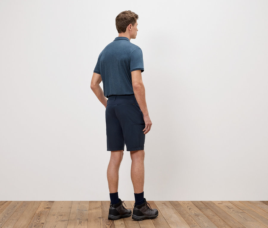 Bir adam sırtı kameraya dönük duruyor, beyaz bir duvarın önünde mavi bir polo tişört, şort ve yürüyüş ayakkabıları giyiyor.