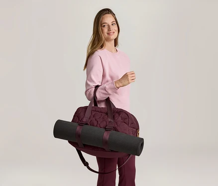 Pembe sweatshirt ve bordo pantolonlu bir kadın, omzunda siyah yoga matı olan bordo bir spor çantası taşıyor.
