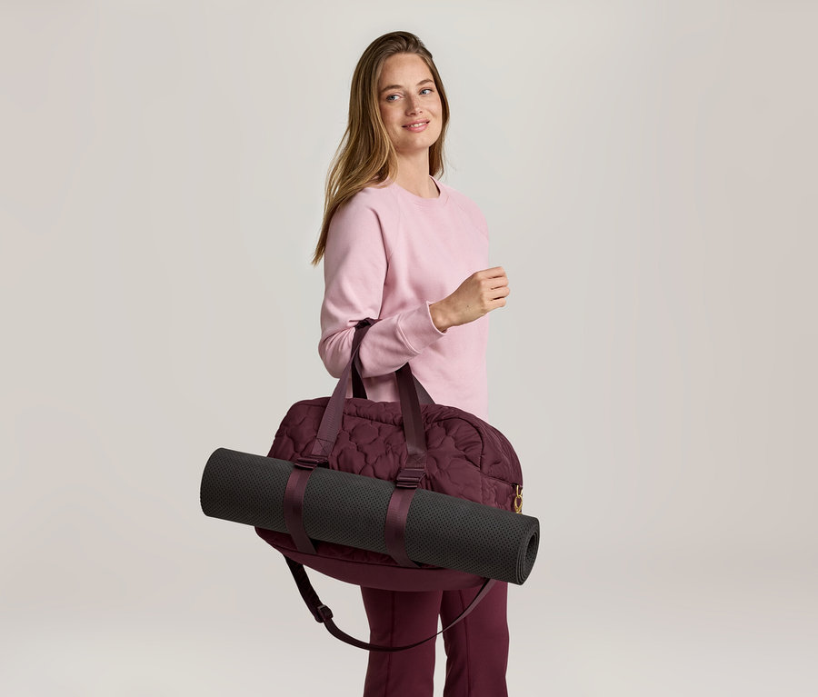 Pembe sweatshirt ve bordo pantolonlu bir kadın, omzunda siyah yoga matı olan bordo bir spor çantası taşıyor.