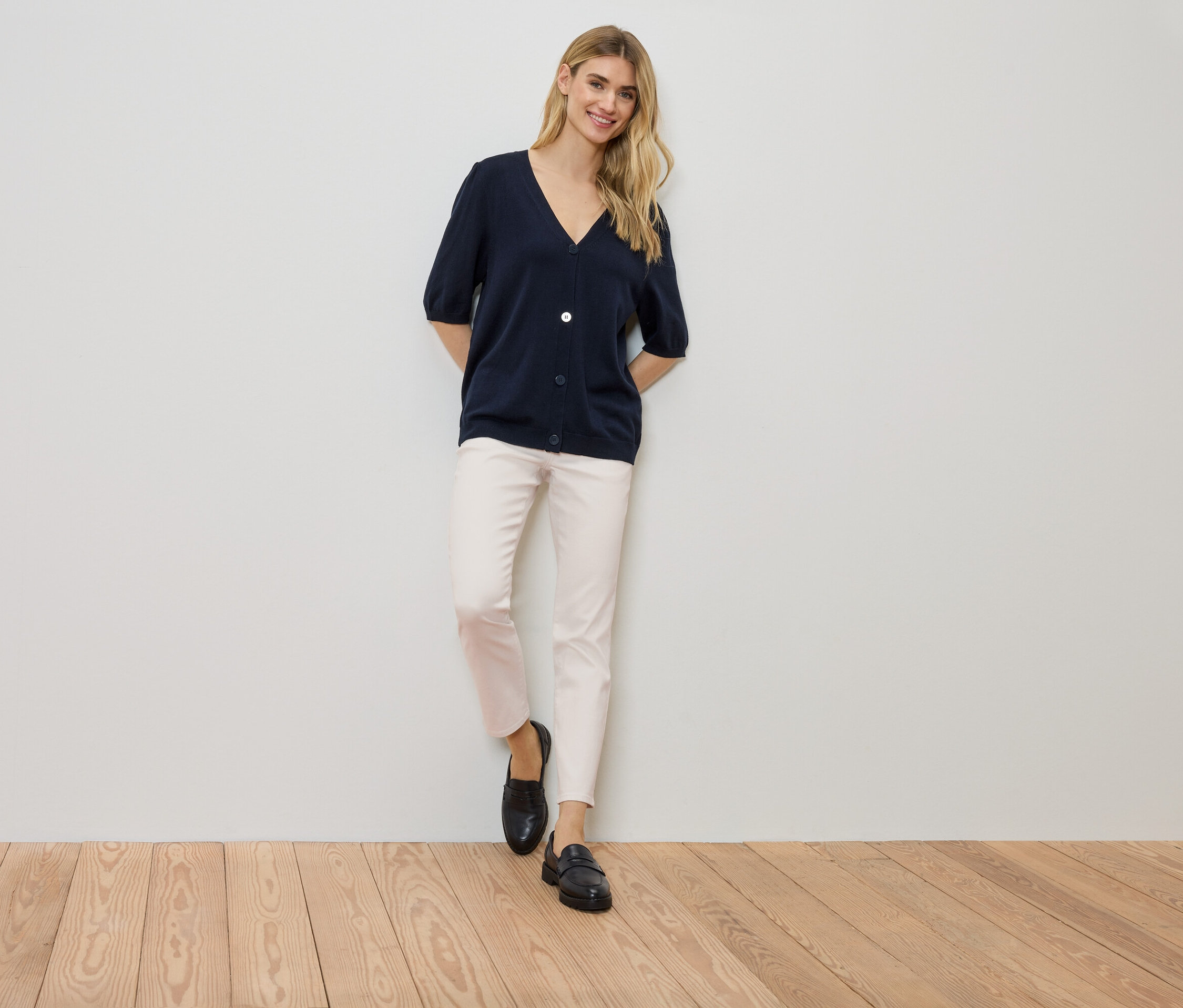 Kadın bir duvara yaslanmış, lacivert bir hırka, açık renkli pantolon ve siyah ayakkabılar giyiyor.