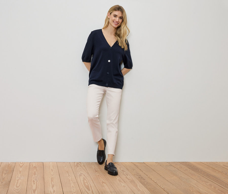 Kadın bir duvara yaslanmış, lacivert bir hırka, açık renkli pantolon ve siyah ayakkabılar giyiyor.